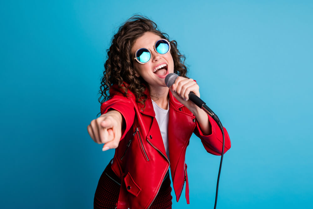 Photo Of Crazy Carefree Lady Open Mouth Sing Hold Mic Direct Finger On You Wear Round Sunglass Isolated On Blue Color Background