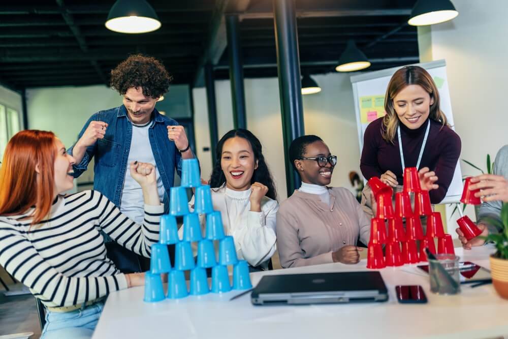 Group Of Young Businesspeople Playing, Building Towers From Plastic Cups, Two Teams, Blue And Red. Teambuilding Activity.