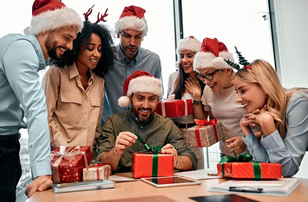 New Year's And Christmas Mood. Group Of Business People In Santa Hats, Colleagues Unpacking Gifts At Work In The Office