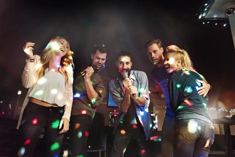 Five friends on a stage singing karaoke, holding microphones, under colorful disco lights and spotlights.