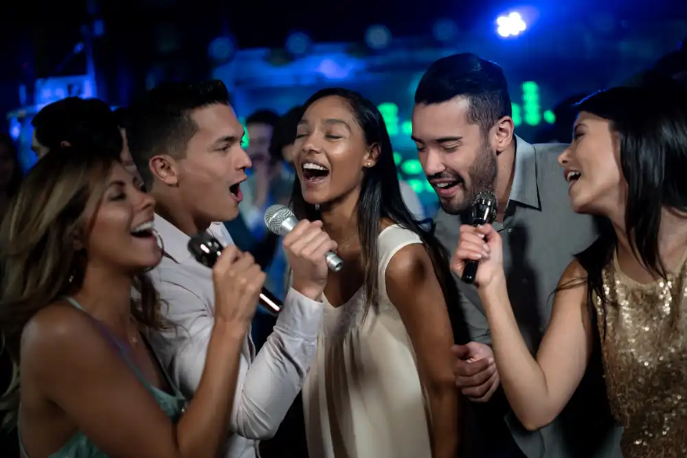 Group of friends singing into microphones at a nightclub, smiling and enjoying a karaoke night on a crowded dance floor in blue lighting.