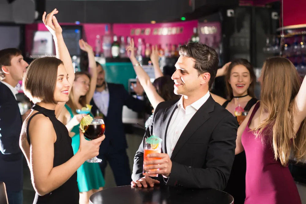 People at a lively bar with cocktails, dancing and raised arms in the background.